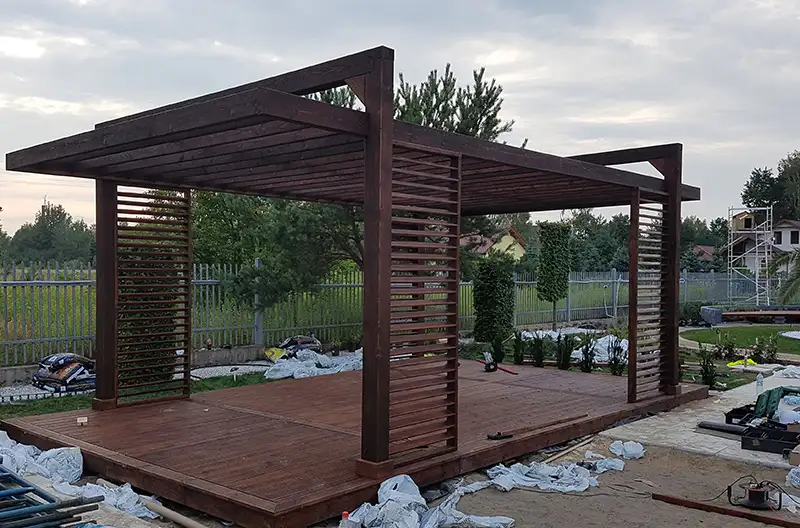 ciemnobrązowa altana ogrodowa na tle drzew stojąca przy płocie