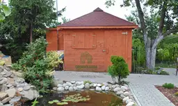 brązowa drewutnia z bordowym dachem stojąca przy drzewie